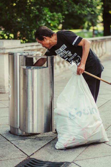 A man with dark hair, wearing a black T-shirt with white graphic text, is seen leaning over a metal trash bin on a paved sidewalk, placing waste into the opening. He holds a large white plastic bag filled with rubbish in his left hand, with the bag partially resting on the ground. The trash bin appears to be made of stainless steel with a shiny, reflective surface and a curved, rounded design. The background includes a stone balustrade, greenery, and trees, suggesting an outdoor setting in a park or urban area. The scene is illuminated by natural daylight, highlighting the details of the bin, the man's attire, and the surrounding environment. This image is related to private waste disposal or rubbish collection services, emphasizing on-site clearance or alternative waste handling options, as managed by Rubbish Clearance Westminster.