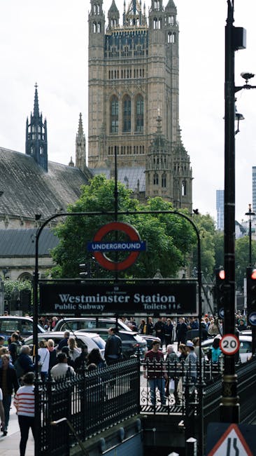 A busy urban scene outside Westminster Station in central London, with a crowd of pedestrians waiting to cross the street and moving along the pavement. In the foreground, a black metal fence runs parallel to the road, separating pedestrians from traffic. To the right, there is a black pole with street signage and a traffic light. Behind the crowd, the station entrance features a black sign with white text reading 'Westminster Station' along with information about public subway toilets. Above, a classic underground roundel sign with a red circle and blue bar reading 'UNDERGROUND' is prominently displayed. In the background, the historic skyline is dominated by the ornate, beige-stone Elizabeth Tower, also known as Big Ben, with its detailed Gothic architecture and tall spires, set against a partly cloudy sky. The environment suggests a typical bustling day in central London, with natural daylight illuminating the scene and highlighting the mixture of historic and modern elements in the area. This scene reflects the vibrant urban setting where independent or private transportation and waste collection services, like those of Rubbish Clearance Westminster, may operate to manage waste from commercial and residential activities nearby.