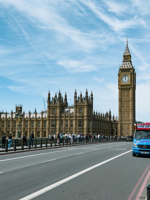 A wide view of the Palace of Westminster in London with its Gothic architecture and multiple spires, including the prominent clock tower known as Big Ben. The scene shows a busy street in the foreground with a line of pedestrians waiting or walking along the pavement, separated from the road by black metal barriers. To the right, there is a parked blue van with partially visible branding, likely a commercial vehicle. The sky above is mostly clear with some thin, wispy clouds, and the lighting suggests daytime with natural sunlight illuminating the detailed stone facade and casting soft shadows. The environment captures a typical sight near a central UK landmark, reflecting the city's bustling urban atmosphere, and it subtly aligns with the concept of private or independent collection services that operate in this busy area of Westminster, away from official waste disposal channels, supporting options like rubbish removal services provided by companies such as Rubbish Clearance Westminster.