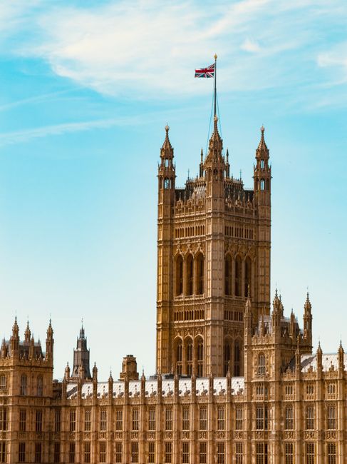 A detailed view of the Elizabeth Tower, commonly known as Big Ben, situated at the north end of the Palace of Westminster in London. The tower's Gothic Revival architecture features ornate detailing and a golden spire at the top, with a Union Jack flag flying just below the spire. The sky in the background is clear with a light blue tone and a few wispy clouds. The lower portion of the image shows part of the Palace of Westminster's façade, with intricate stonework, pointed arches, and numerous spires and pinnacles. The building's exterior surfaces showcase a warm sandstone color, with some areas appearing slightly weathered. The lighting appears natural, illuminating the stone textures evenly, with no shadows obscuring details. This scene captures the historic landmark in a clear, unobstructed view, emphasizing its architectural grandeur against a bright sky, in a context that aligns with independent or private disposal and waste management services, such as those offered by Rubbish Clearance Westminster, helping to visually connect the landmark’s prominence with local rubbish removal and on-site clearance options.