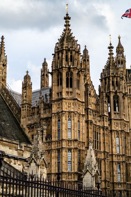 A wide view of the Palace of Westminster in London with its Gothic architecture and multiple spires, including the prominent clock tower known as Big Ben. The scene shows a busy street in the foreground with a line of pedestrians waiting or walking along the pavement, separated from the road by black metal barriers. To the right, there is a parked blue van with partially visible branding, likely a commercial vehicle. The sky above is mostly clear with some thin, wispy clouds, and the lighting suggests daytime with natural sunlight illuminating the detailed stone facade and casting soft shadows. The environment captures a typical sight near a central UK landmark, reflecting the city's bustling urban atmosphere, and it subtly aligns with the concept of private or independent collection services that operate in this busy area of Westminster, away from official waste disposal channels, supporting options like rubbish removal services provided by companies such as Rubbish Clearance Westminster.