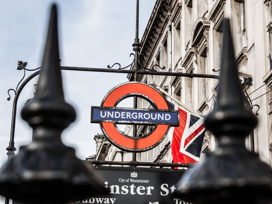 A close-up photograph taken from behind black metal fence spikes shows a London Underground roundel sign with a red circle and blue bar displaying the word 'UNDERGROUND' in white text. The sign hangs from a decorative wrought iron arm attached to an ornate building facade with detailed stonework and large windows. Behind the sign, a British flag is partially visible, fluttering against a cloudy sky. The scene suggests an urban environment typical of central London, where private waste collection services like Rubbish Clearance Westminster may operate in proximity to prominent public transport hubs, supporting alternative rubbish removal options outside of municipal waste rules. The building's architectural style and the signage evoke a historic yet busy city atmosphere, emphasizing the importance of proper waste management in such dense and structured urban settings.