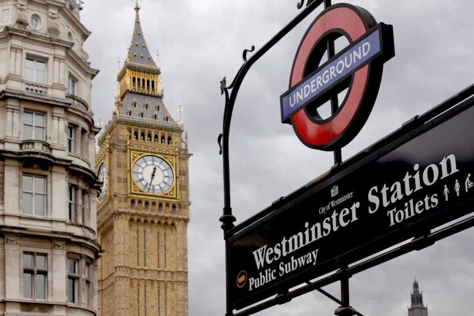 A close-up view of the Westminster underground station sign, featuring the classic red roundel with a blue bar displaying the word 'UNDERGROUND' in white text. Positioned above the sign is a decorative black metal arch with scrolling details. Behind the sign, the iconic Big Ben clock tower is visible, showcasing its ornate Gothic Revival architecture with a gold and stone façade, a large clock face displaying the time, and a pointed spire topped with a small flag. To the left, a segment of a curved, light-colored building with multiple windows and decorative cornices can be seen, illustrating typical London architecture. The sky is overcast with grey clouds, creating a subdued atmosphere. This scene reflects a central London setting where public transport infrastructure and historic landmarks coexist, illustrating the urban environment that a rubbish removal service like Rubbish Clearance Westminster might operate within, often handling waste from such busy city areas or nearby developments requiring professional waste management and alternative disposal solutions rather than local authority collection.