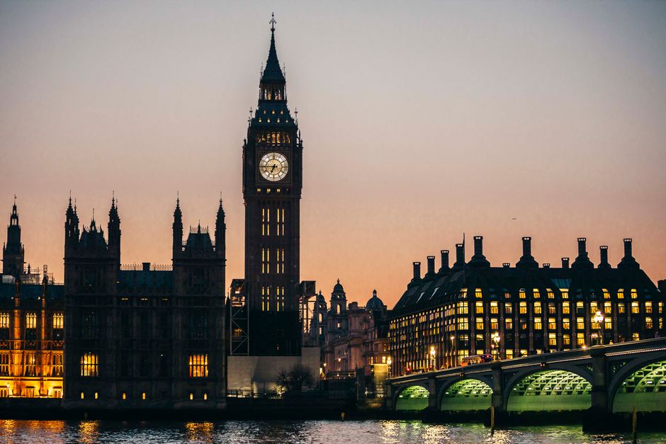 A night-time view of London's iconic Big Ben clock tower and the Houses of Parliament, illuminated with warm yellow lighting from many windows and exterior lights, with a bridge crossing the River Thames in the foreground. The sky is dusky, with a faint gradient from pale pink to darker blue. The buildings are detailed with Gothic architectural elements, pinnacles, and spires, and reflect softly on the water below. No rubbish or waste materials are visible in the image. This scenic cityscape captures a well-lit, historic part of Westminster, providing a professional and neutral visual context unrelated to rubbish removal services.