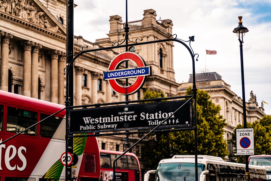 A close-up view of the Westminster underground station sign, featuring the classic red roundel with a blue bar displaying the word 'UNDERGROUND' in white text. Positioned above the sign is a decorative black metal arch with scrolling details. Behind the sign, the iconic Big Ben clock tower is visible, showcasing its ornate Gothic Revival architecture with a gold and stone façade, a large clock face displaying the time, and a pointed spire topped with a small flag. To the left, a segment of a curved, light-colored building with multiple windows and decorative cornices can be seen, illustrating typical London architecture. The sky is overcast with grey clouds, creating a subdued atmosphere. This scene reflects a central London setting where public transport infrastructure and historic landmarks coexist, illustrating the urban environment that a rubbish removal service like Rubbish Clearance Westminster might operate within, often handling waste from such busy city areas or nearby developments requiring professional waste management and alternative disposal solutions rather than local authority collection.